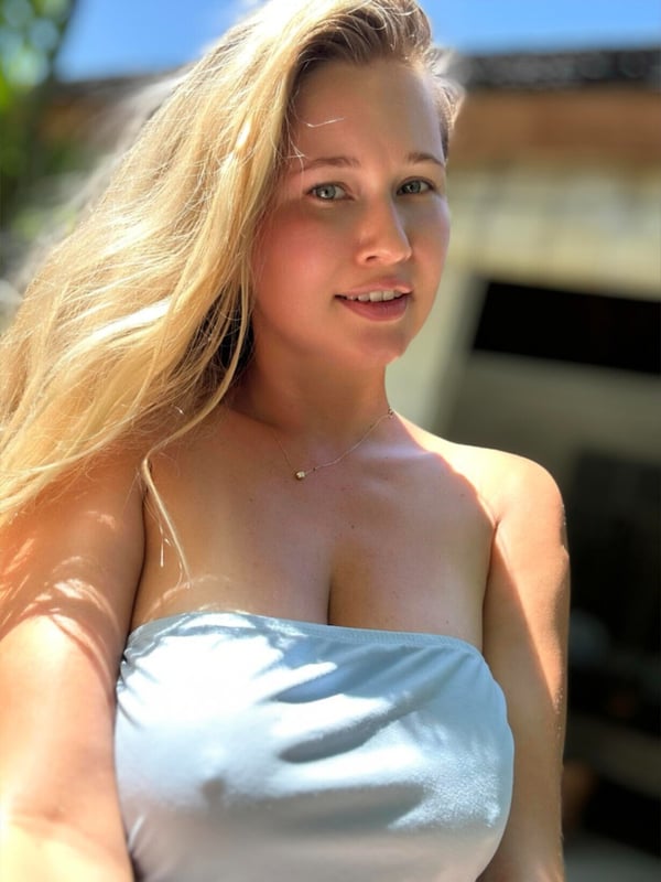 Young woman with long blonde hair smiling at the camera, wearing a light blue strapless top, standing outdoors in sunlight near tropical surroundings.