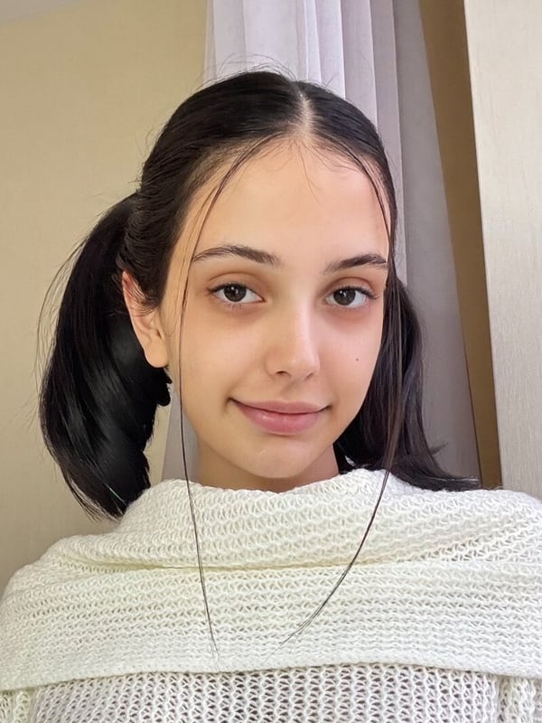 Close-up portrait of a young woman with dark hair in pigtails, wearing a white knit top, smiling gently indoors.