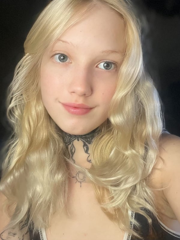 Close-up portrait of a young woman with long blonde wavy hair, choker necklace, neck tattoos, and green eyes, smiling gently in a dimly lit room.