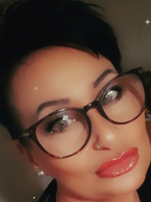 Woman with short dark hair and glasses, smiling with glossy lips in a close-up indoor selfie with decorative background elements.