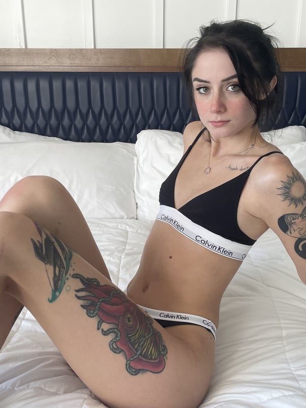 A woman posing on a bed in a white Calvin Klein bra and panties, displaying tattoos on her arms and legs.