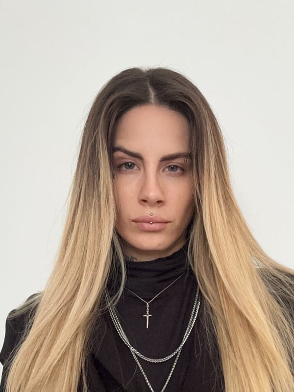 Close-up portrait of a woman with long blonde hair, wearing a black turtleneck and layered necklaces, gazing directly at the camera against a white background.