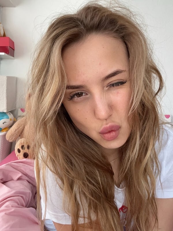 A young woman with long wavy hair taking a selfie in a bedroom setting.
