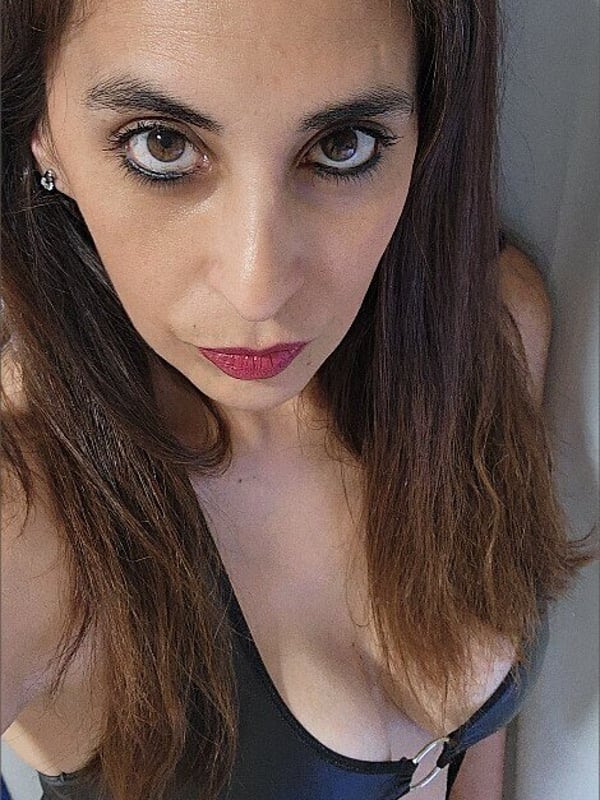 Close-up portrait of a woman with long dark hair and striking makeup, wearing a shiny black top, gazing intensely at the camera.