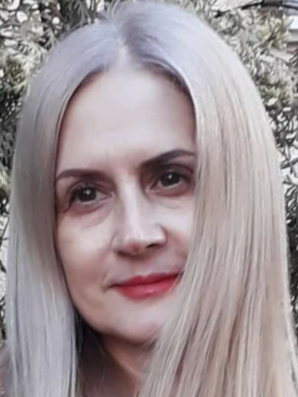 Close-up portrait of a woman with long silver-blonde hair and red lipstick, smiling softly against a blurred outdoor background with greenery.