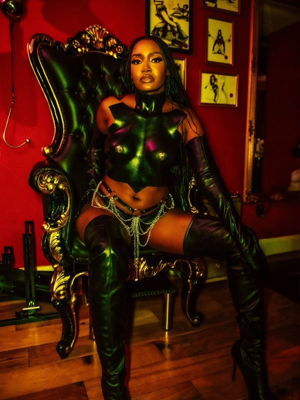 A woman seated confidently on an ornate chair in a dimly lit room, dressed in sleek black leather outfit with long gloves and boots.