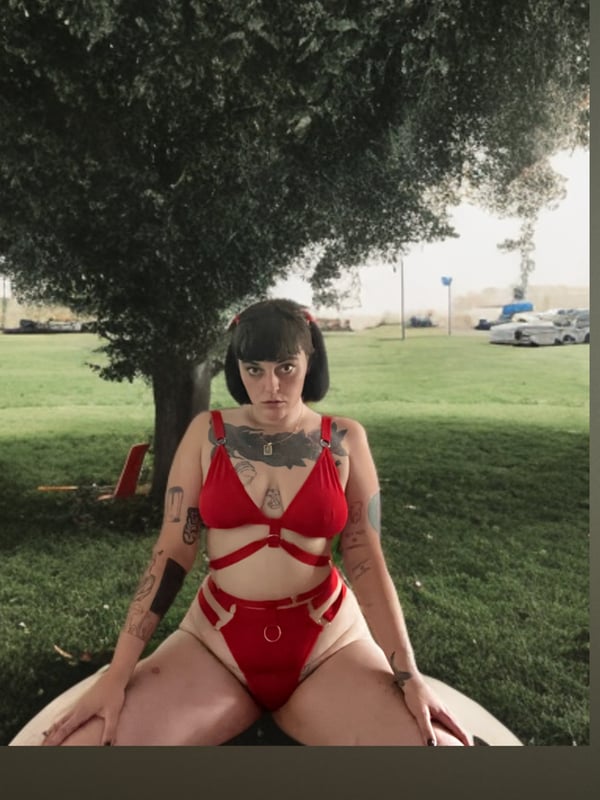 A woman with tattoos and dark bangs sitting outdoors under a tree in a red harness-style outfit on the grass.