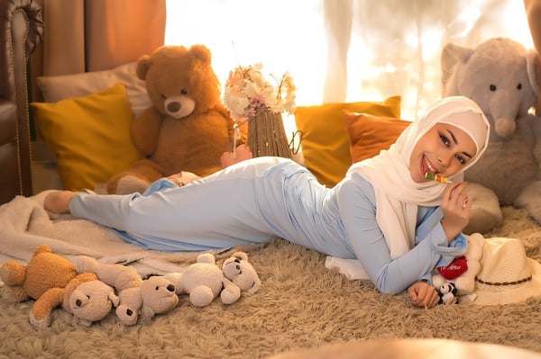 Young woman in a light blue outfit and white hijab lounging playfully on a rug surrounded by teddy bears and pillows in a warmly lit room.