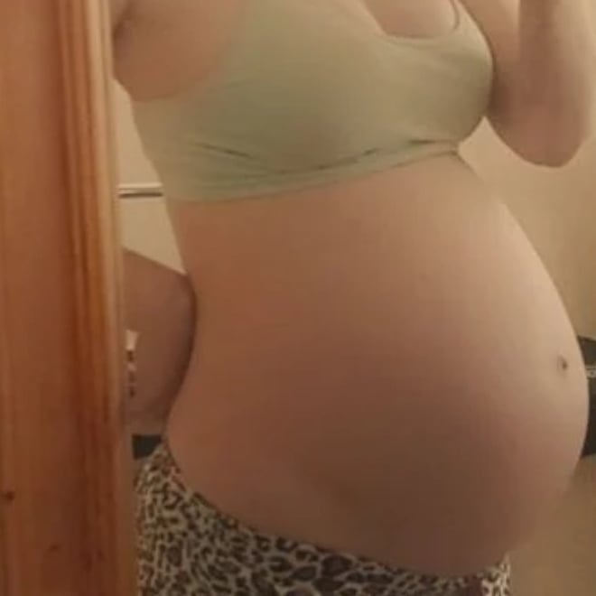 A pregnant woman standing in front of a mirror, wearing a light-colored top and patterned bottoms, posing with her hand on her hip.