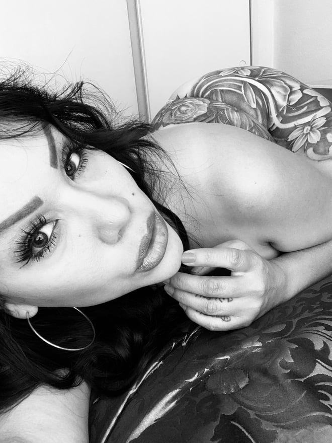 A woman with long dark hair and tattoos poses reclined on a bed in black and white, wearing a patterned top.