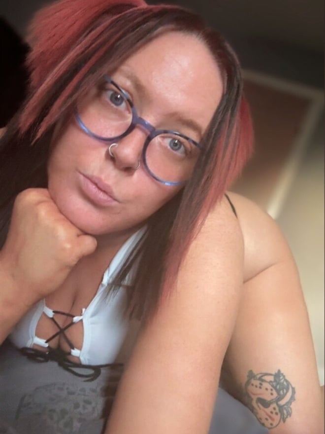 Woman with red hair and glasses wearing a white top, posing with hand on chin indoors.