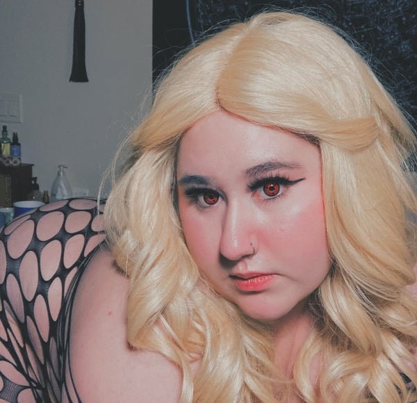 A woman with long blonde hair and dramatic makeup posing in a dimly lit room with hanging decor and shelves in the background.