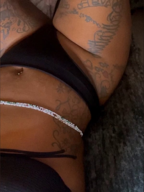 Close-up of a woman in a black bikini top, displaying arm tattoos and a belly chain.