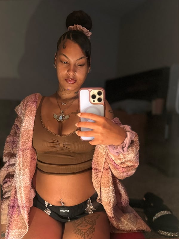 A woman with a bun hairstyle sitting on a bed, wearing a fuzzy robe over a top and shorts, holding her phone with jewelry and tattoos visible.