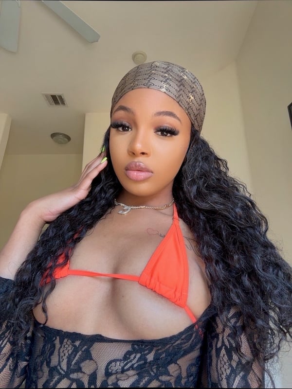 A young woman with long wavy black hair wearing a patterned headwrap, gold jewelry, a pink triangle bikini top, and black lace top, posing confidently indoors.