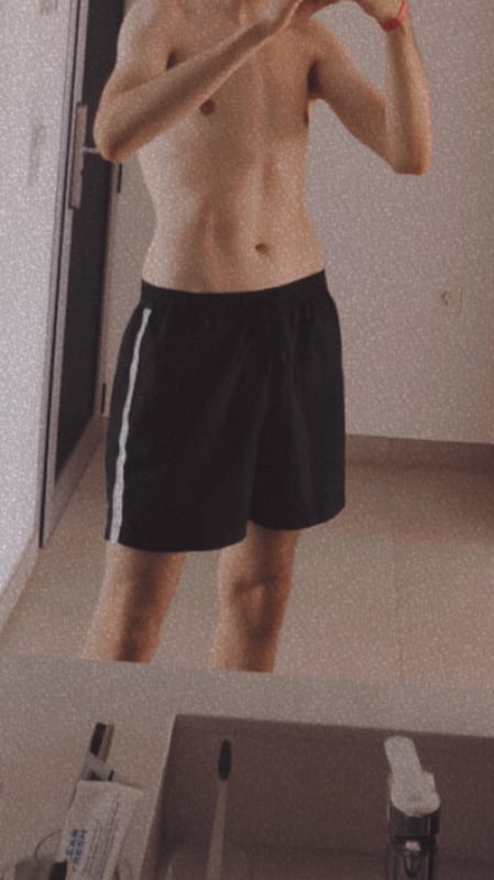 Young man taking a mirror selfie in shorts, standing in a bathroom with a neutral background.