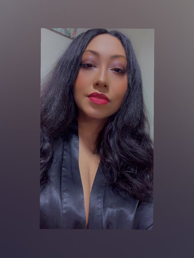 Woman with long dark hair and red lipstick, posing confidently in a low-cut black satin top indoors.