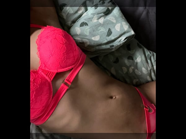A woman lying on a bed wearing a pink lace bra and green patterned clothing, posing relaxedly indoors.