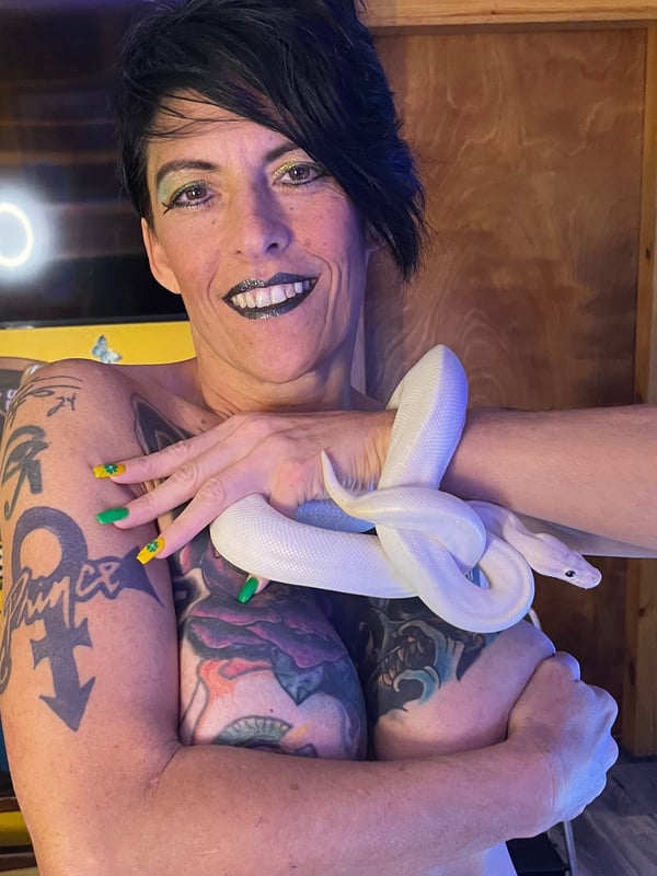 A woman with short dark hair and tattoos holds a white snake draped over her shoulders in a home setting.