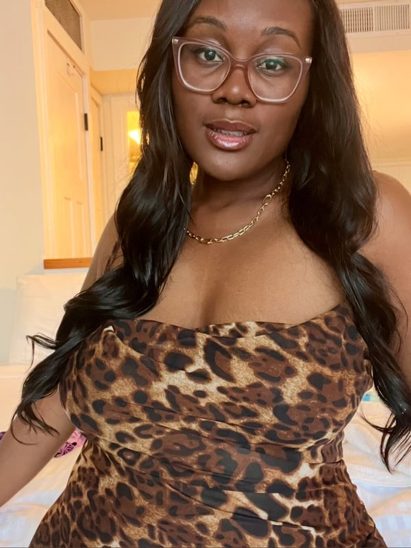 Young woman with glasses and long dark hair smiling while sitting on a bed in a leopard print top inside a bedroom.