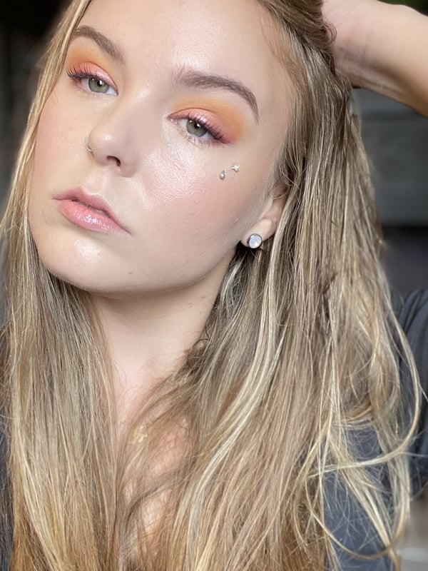 A young woman with long blonde hair and orange eyeshadow posing confidently indoors, wearing a dark top.
