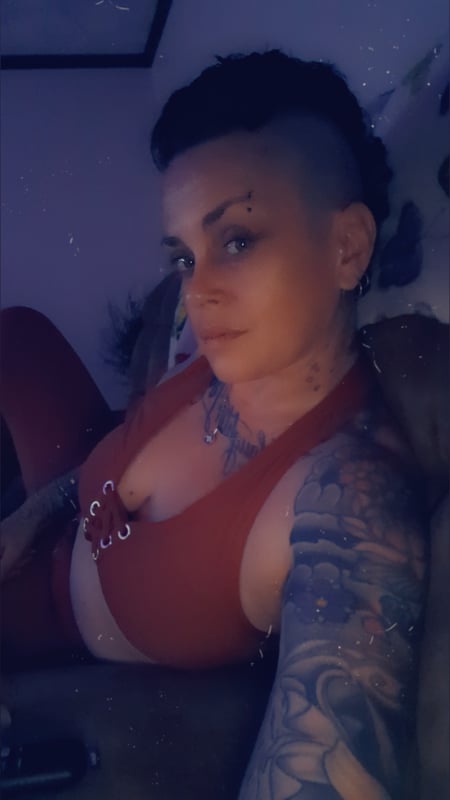A woman with a mohawk hairstyle and tattoos poses confidently indoors, wearing a red top and leggings while lounging on furniture.
