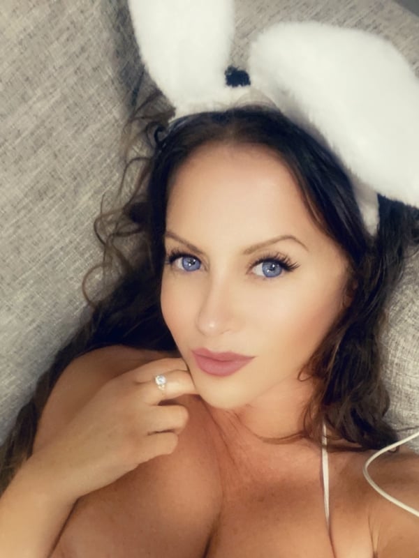 A woman with long wavy hair and blue eyes wearing white bunny ears, posing playfully on a couch.