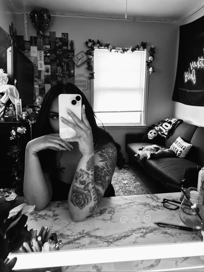 Woman with long dark hair and tattoos on her arm taking a mirror selfie in a cozy room with decorations and a couch.