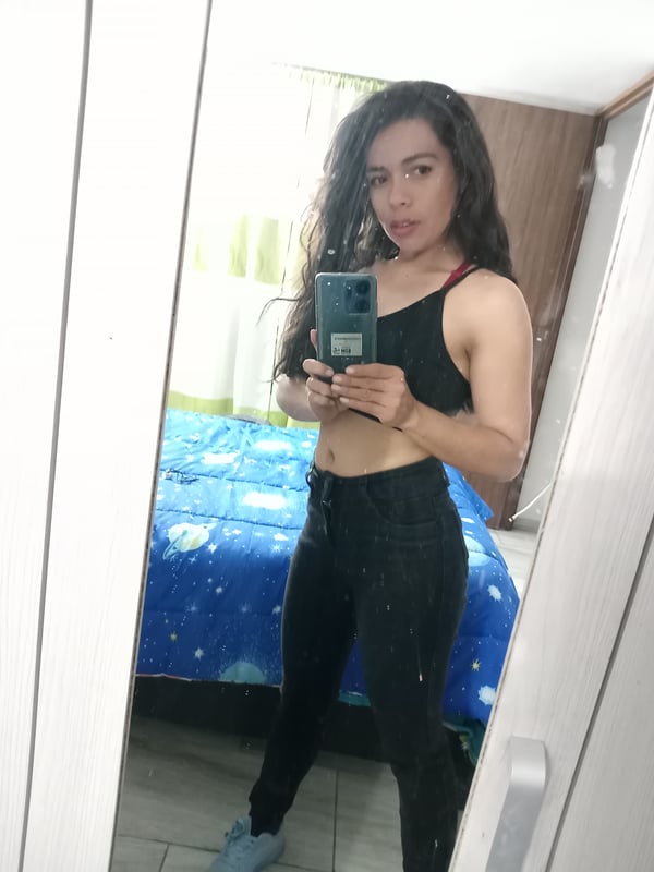 Young woman taking a mirror selfie in a black tank top and leggings, standing in a bedroom with a blue bedspread.
