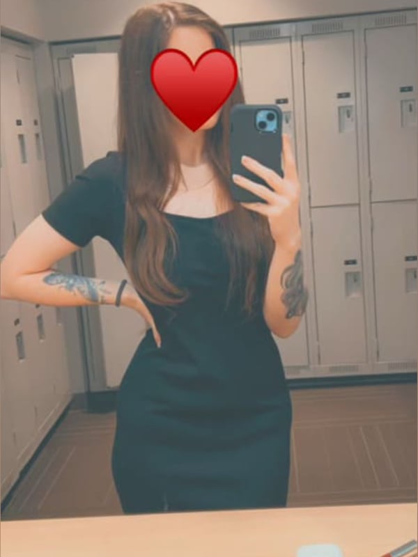 A woman with long dark hair taking a selfie in a black dress inside a locker room, heart emoji over her face, tattoos on her arms.