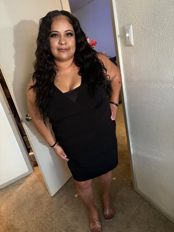 A woman with long wavy dark hair posing confidently in a black dress inside a home near an open door.