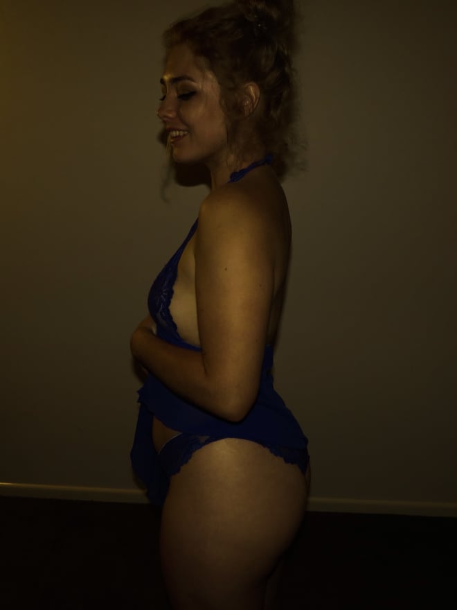 A young woman with curly hair stands sideways in a blue lingerie top and bottoms, posing confidently in a neutral indoor setting.