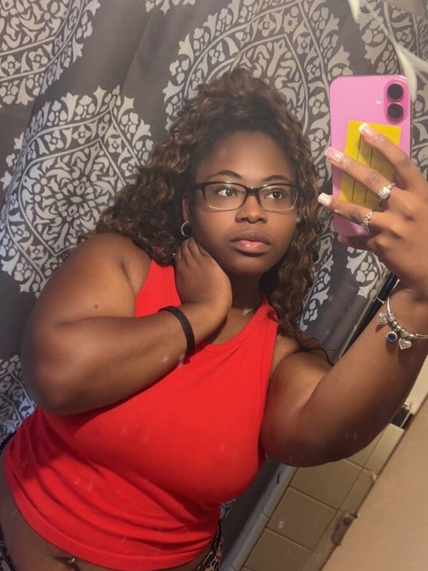 A woman taking a selfie in a bathroom, wearing glasses and a red tank top, posing confidently with her hand on her neck.
