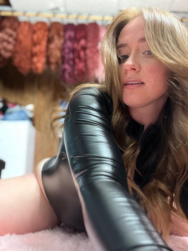 A woman with long blonde hair smiling softly while wearing a shiny black leather outfit, posed against a background of colorful hanging fabrics.