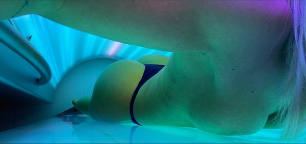 A person lying on their stomach in a tanning bed with blue glowing lights, wearing dark bottoms.