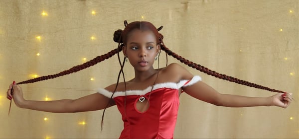 A woman with long braided hair wearing a red off-shoulder dress poses dynamically with arms extended against a sparkling golden backdrop.