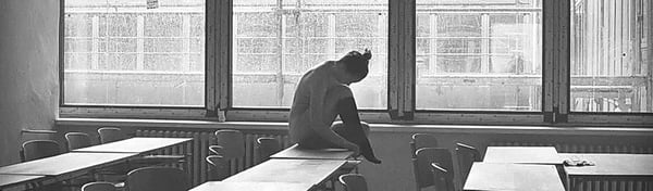A person sits pensively by a window in a classroom with desks, captured in black and white photography.