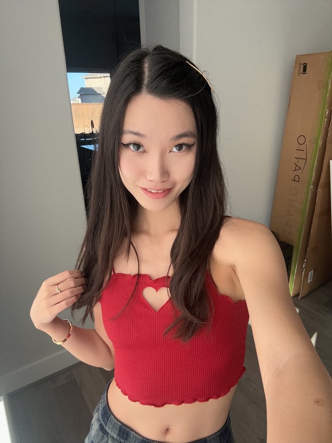 A woman with long dark hair smiling at the camera while wearing a red cropped top and jeans indoors.