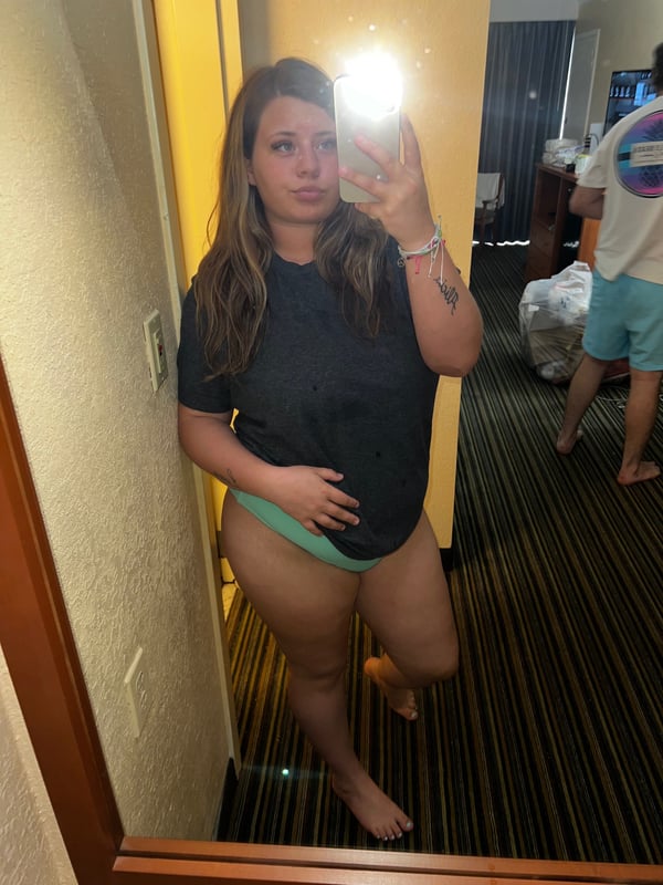 A woman taking a mirror selfie in a hotel room, dressed in a dark t-shirt and light green underwear, standing near a yellow wall.