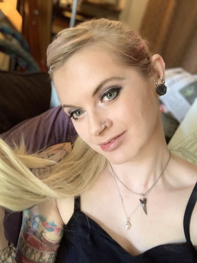 Close-up portrait of a blonde woman with piercings, tattoos, and green eyes sitting indoors.