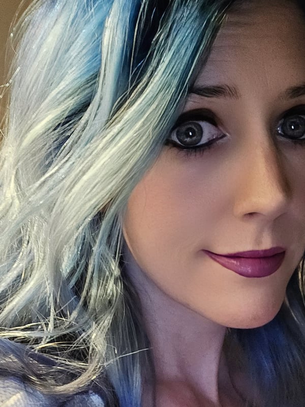 Close-up portrait of a woman with long dyed blue-tinted blonde hair, striking blue eyes, and a subtle smile.
