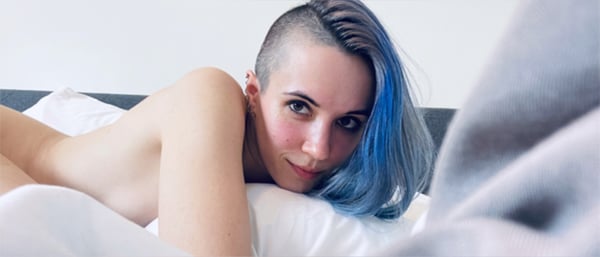 A young woman with blue hair reclining on a bed, smiling at the camera in a relaxed pose.