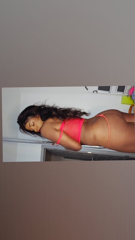 A woman posing sideways on a bed in a pink bikini bottom and top, with long dark hair and a relaxed indoor setting.