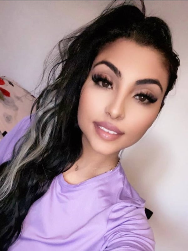 A woman with wavy dark hair streaked with lighter highlights, wearing a purple top, smiling at the camera in a close-up selfie.