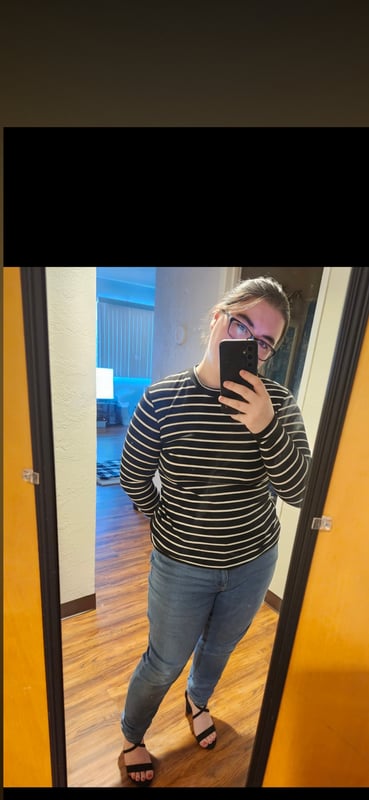 Young woman with glasses taking a selfie in a bathroom mirror, wearing a striped shirt and jeans.