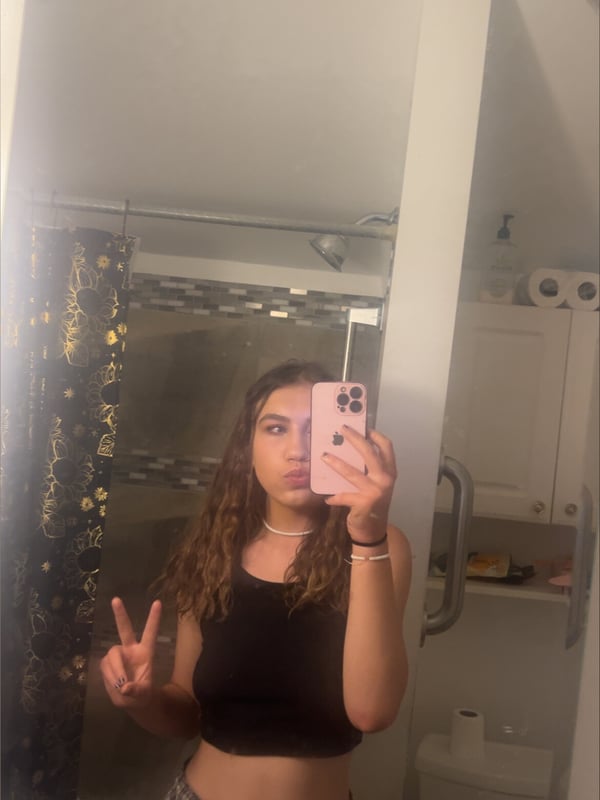 Young woman with wavy hair making a peace sign in front of a bathroom mirror, dressed in a black crop top and checkered pants.