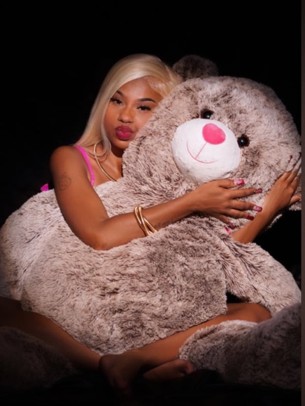 A woman with blonde hair smiling while hugging a large teddy bear in a dimly lit setting.