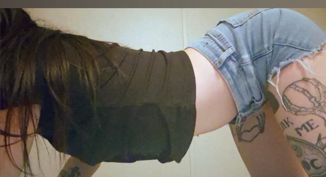 A young woman with long dark hair poses playfully on the floor in a casual top and denim shorts, with visible tattoos on her arms and legs.
