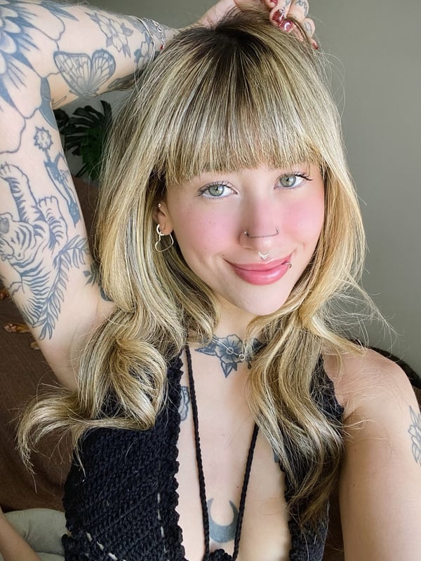 Close-up of a smiling young woman with blonde hair, tattoos on arms and neck, wearing a black top, posing confidently indoors.