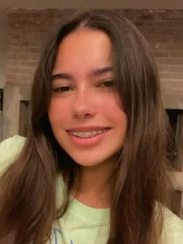 A young woman with long brown hair and braces smiling at the camera indoors.